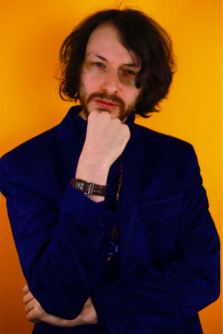 Jump tag Club founder Rito with messy hair in a blue suit against an orange background, hand touching his chin